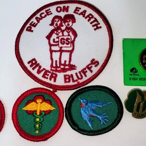 K&ouml;nnte beinhalten: Eine Sammlung von Girl Scout-Abzeichen und Anstecknadeln. Die Abzeichen umfassen einen gro&szlig;en runden Aufn&auml;her mit den Worten "Peace on Earth" und "River Bluffs" sowie kleinere Aufn&auml;her mit verschiedenen Designs. Es gibt auch eine Anstecknadel auf einer gr&uuml;nen Karte.