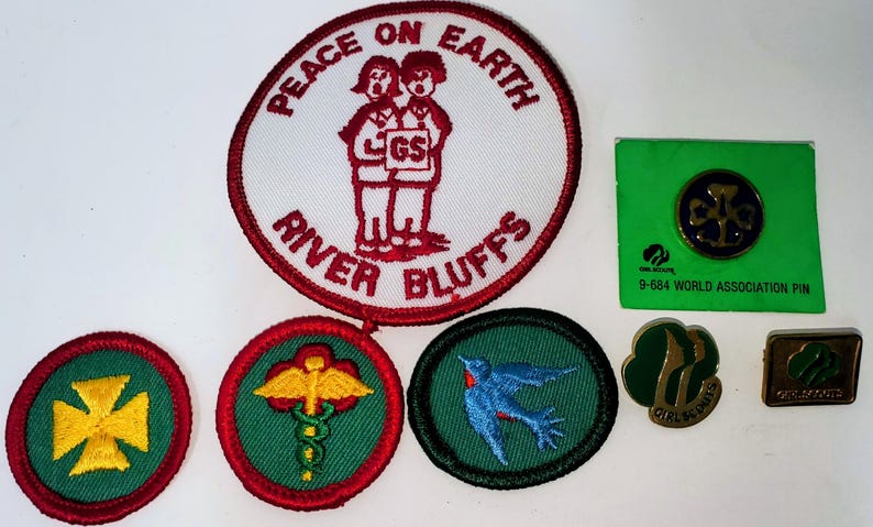 K&ouml;nnte beinhalten: Eine Sammlung von Girl Scout-Abzeichen und Anstecknadeln. Die Abzeichen umfassen einen gro&szlig;en runden Aufn&auml;her mit den Worten "Peace on Earth" und "River Bluffs" sowie kleinere Aufn&auml;her mit verschiedenen Designs. Es gibt auch eine Anstecknadel auf einer gr&uuml;nen Karte.