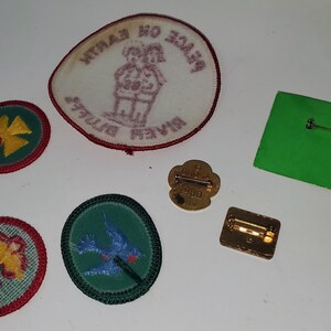 K&ouml;nnte beinhalten: Eine Sammlung von Vintage-Pfadfinderinnen-Abzeichen und -Anstecknadeln. Die Aufn&auml;her zeigen gestickte Designs eines vierbl&auml;ttrigen Kleeblatts, einer Blume und eines blauen Vogels. Es gibt auch einen Aufn&auml;her mit zwei Figuren und Text. Die Anstecknadeln sind goldfarben.