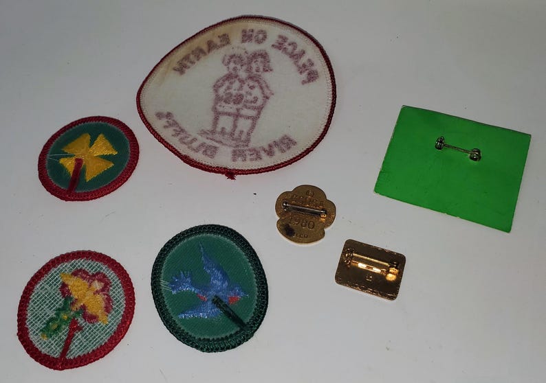 K&ouml;nnte beinhalten: Eine Sammlung von Vintage-Pfadfinderinnen-Abzeichen und -Anstecknadeln. Die Aufn&auml;her zeigen gestickte Designs eines vierbl&auml;ttrigen Kleeblatts, einer Blume und eines blauen Vogels. Es gibt auch einen Aufn&auml;her mit zwei Figuren und Text. Die Anstecknadeln sind goldfarben.
