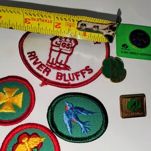 K&ouml;nnte beinhalten: Eine Sammlung von Pfadfinderinnen-Abzeichen und Anstecknadeln. Enth&auml;lt einen River Bluffs-Aufn&auml;her, einen blauen Vogel-Aufn&auml;her, einen goldenen Kreuz-Aufn&auml;her und eine gr&uuml;n-goldene Anstecknadel. Ein gelbes Ma&szlig;band ist im Hintergrund.