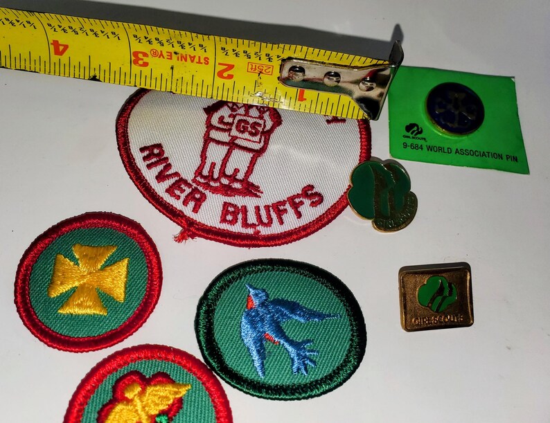 K&ouml;nnte beinhalten: Eine Sammlung von Pfadfinderinnen-Abzeichen und Anstecknadeln. Enth&auml;lt einen River Bluffs-Aufn&auml;her, einen blauen Vogel-Aufn&auml;her, einen goldenen Kreuz-Aufn&auml;her und eine gr&uuml;n-goldene Anstecknadel. Ein gelbes Ma&szlig;band ist im Hintergrund.