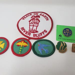 K&ouml;nnte beinhalten: Eine Sammlung von Pfadfinderinnen-Abzeichen und Anstecknadeln. Die Abzeichen umfassen ein gro&szlig;es wei&szlig;es und rotes Abzeichen mit den Worten "Peace on Earth" und "River Bluffs". Andere Abzeichen zeigen ein gelbes Kreuz, einen Caduceus und einen blauen Vogel. Es gibt auch zwei Anstecknadeln.