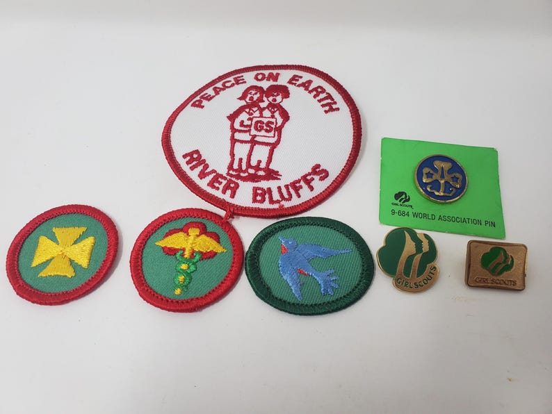 K&ouml;nnte beinhalten: Eine Sammlung von Pfadfinderinnen-Abzeichen und Anstecknadeln. Die Abzeichen umfassen ein gro&szlig;es wei&szlig;es und rotes Abzeichen mit den Worten "Peace on Earth" und "River Bluffs". Andere Abzeichen zeigen ein gelbes Kreuz, einen Caduceus und einen blauen Vogel. Es gibt auch zwei Anstecknadeln.