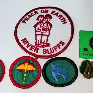 K&ouml;nnte beinhalten: Eine Sammlung von Girl Scout-Aufn&auml;hern und -Anstecknadeln. Die Aufn&auml;her umfassen einen gro&szlig;en runden Aufn&auml;her mit dem Text "PEACE ON EARTH RIVER BLUFFS" und einem Design von zwei Figuren. Andere Aufn&auml;her und Anstecknadeln zeigen verschiedene Designs und Logos.