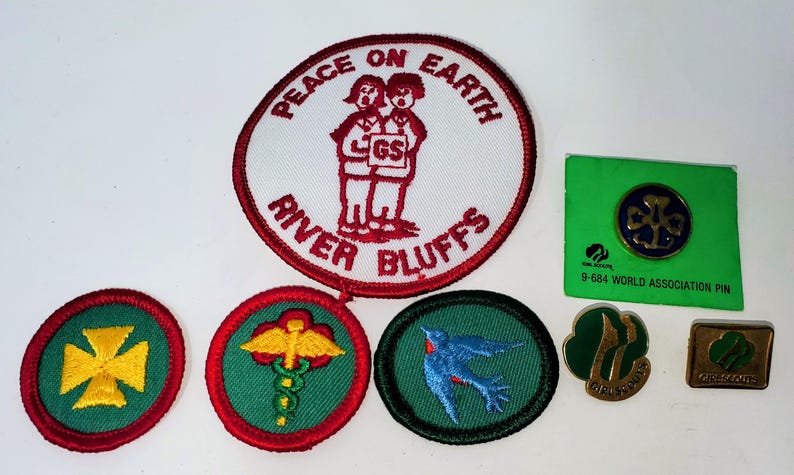 K&ouml;nnte beinhalten: Eine Sammlung von Girl Scout-Aufn&auml;hern und -Anstecknadeln. Die Aufn&auml;her umfassen einen gro&szlig;en runden Aufn&auml;her mit dem Text "PEACE ON EARTH RIVER BLUFFS" und einem Design von zwei Figuren. Andere Aufn&auml;her und Anstecknadeln zeigen verschiedene Designs und Logos.
