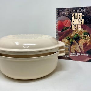 Puede incluir: Un juego de cocina apilable para microondas de plástico color crema con tapa a juego, acompañado de un libro de cocina de espiral titulado TUPPERWAVE STACK-COOKED MEALS, que presenta una fotografía de un plato de pasta en la portada.