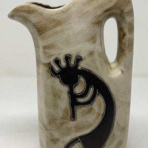 May include: A ceramic pitcher with a beige and brown mottled finish. A black Kokopelli figure is featured on the front. The pitcher has a handle and a spout.