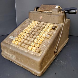 May include: A vintage brown metal adding machine with a cream-colored keyboard and a hand crank. The machine has a paper roll holder and a small display window.