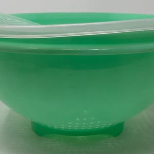 May include: A bright mint green plastic colander bowl featuring a wide rim and a translucent white lid. The base of the bowl has a series of small drainage holes arranged in a circular pattern, and the structure is supported by a raised pedestal base.