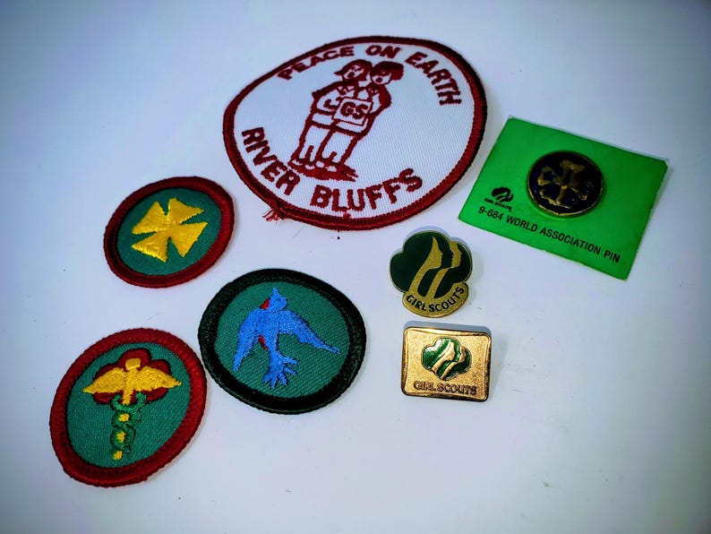 K&ouml;nnte beinhalten: Eine Sammlung von Pfadfinderinnen-Abzeichen und Anstecknadeln. Die Abzeichen umfassen ein rot-wei&szlig;es Abzeichen mit den Worten "Peace on Earth" und "River Bluffs". Andere Abzeichen zeigen ein Kreuz, einen blauen Vogel und ein medizinisches Symbol. Es gibt auch Pfadfinderinnen-Anstecknadeln.