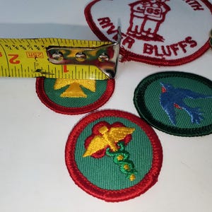K&ouml;nnte beinhalten: Eine Sammlung von Pfadfinderinnen-Abzeichen und Anstecknadeln. Die gestickten Abzeichen zeigen verschiedene Designs, darunter einen Vogel, ein gelbes Symbol und ein medizinisches Symbol. Die Abzeichen haben einen Durchmesser von ca. 5 cm, gemessen mit einem Ma&szlig;band.