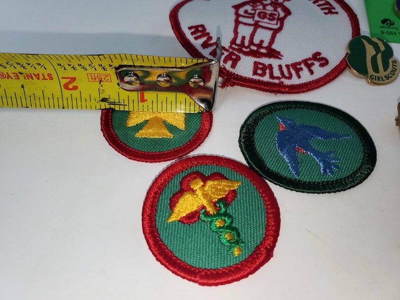 K&ouml;nnte beinhalten: Eine Sammlung von Pfadfinderinnen-Abzeichen und Anstecknadeln. Die gestickten Abzeichen zeigen verschiedene Designs, darunter einen Vogel, ein gelbes Symbol und ein medizinisches Symbol. Die Abzeichen haben einen Durchmesser von ca. 5 cm, gemessen mit einem Ma&szlig;band.