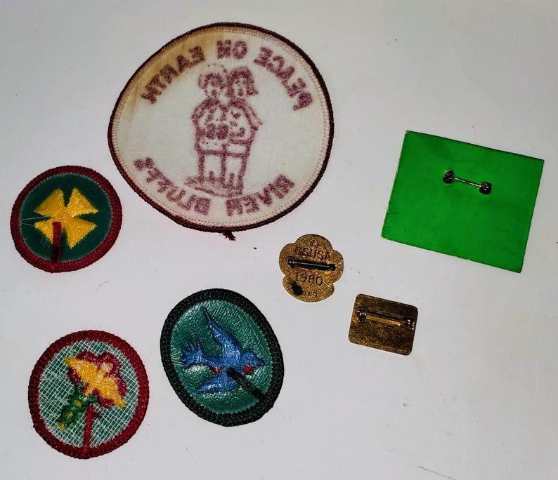 K&ouml;nnte beinhalten: Eine Sammlung von Vintage-Aufn&auml;hern und Anstecknadeln, darunter eine gr&uuml;ne quadratische Anstecknadel, eine goldene Anstecknadel mit "GSA USA 1980" und mehrere gestickte Aufn&auml;her mit Blumen- und Vogelmotiven. Ein Aufn&auml;her lautet "Peace on Earth River Bluffs".