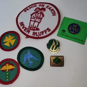 K&ouml;nnte beinhalten: Eine Sammlung von Girl Scout-Abzeichen und -Anstecknadeln. Enth&auml;lt einen runden Aufn&auml;her mit dem Text "Peace on Earth" und "River Bluffs", eine gr&uuml;ne Anstecknadel mit einem blauen Emblem und mehrere andere Aufn&auml;her mit verschiedenen Designs und Farben.