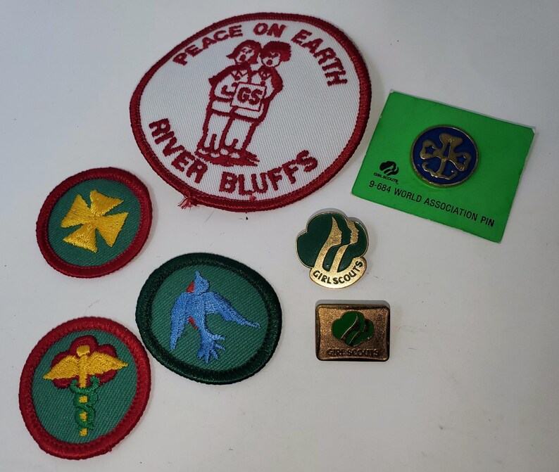 K&ouml;nnte beinhalten: Eine Sammlung von Girl Scout-Abzeichen und -Anstecknadeln. Enth&auml;lt einen runden Aufn&auml;her mit dem Text "Peace on Earth" und "River Bluffs", eine gr&uuml;ne Anstecknadel mit einem blauen Emblem und mehrere andere Aufn&auml;her mit verschiedenen Designs und Farben.