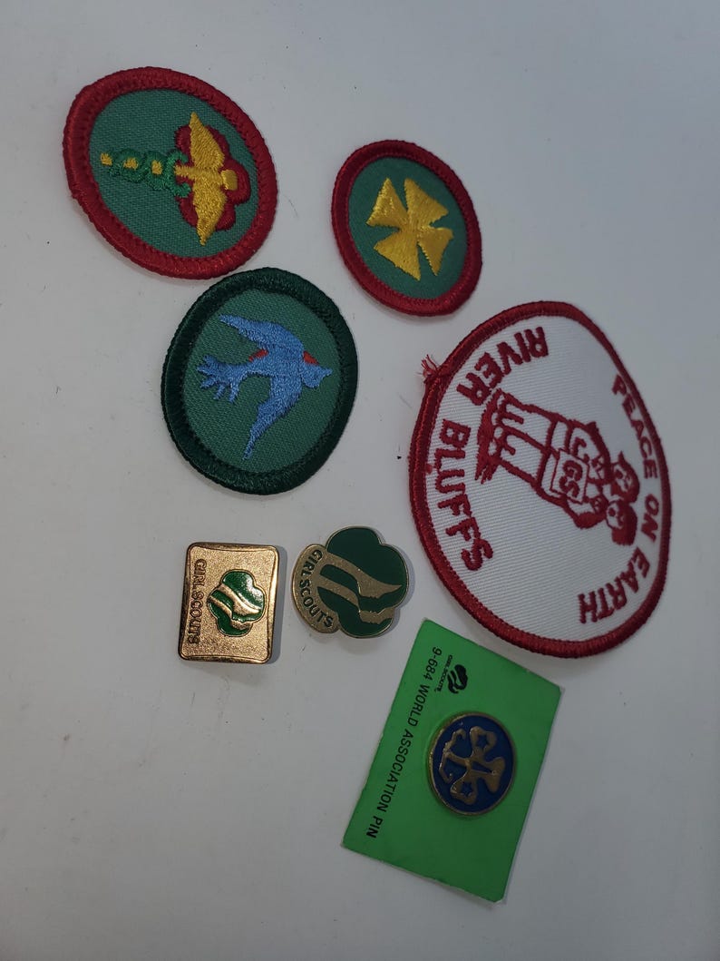 K&ouml;nnte beinhalten: Eine Sammlung von Pfadfinderinnen-Abzeichen und Anstecknadeln. Die Abzeichen zeigen verschiedene Designs, darunter einen Vogel, ein vierbl&auml;ttriges Kleeblatt und den Text "River Bluffs" und "Peace on Earth". Die Anstecknadeln umfassen eine goldene quadratische Anstecknadel und eine gr&uuml;ne Kleeblatt-Anstecknadel.
