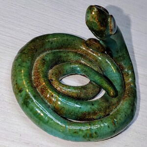 May include: A green ceramic snake sculpture with a textured surface. The snake is coiled in a circle with its head raised.