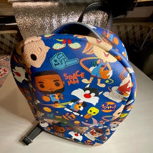 May include: A blue backpack with a cartoon print featuring characters from Space Jam and Looney Tunes. The print includes Bugs Bunny, Tweety Bird, Sylvester, Daffy Duck, Taz, and more. The backpack has a black adjustable strap.