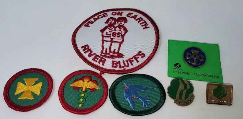 K&ouml;nnte beinhalten: Eine Sammlung von Pfadfinderinnen-Abzeichen und Anstecknadeln. Die Abzeichen umfassen einen gro&szlig;en runden Aufn&auml;her mit den Worten "Peace on Earth" und "River Bluffs" sowie mehrere kleinere runde Aufn&auml;her mit verschiedenen Designs. Es gibt auch mehrere Anstecknadeln.