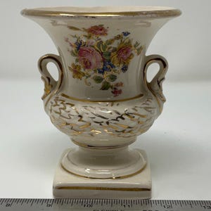 May include: A small, white ceramic urn with gold accents and a floral design. The urn has two handles and a square base.