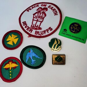 K&ouml;nnte beinhalten: Eine Sammlung von Pfadfinderinnen-Abzeichen und -Anstecknadeln. Die Abzeichen zeigen verschiedene Designs, darunter ein Kreuz, ein Vogel und die Worte &bdquo;Peace on Earth&ldquo; und &bdquo;River Bluffs&ldquo;. Die Anstecknadeln umfassen eine Weltverbandsnadel und ein Pfadfinderinnen-Logo.