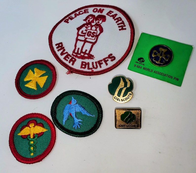 K&ouml;nnte beinhalten: Eine Sammlung von Pfadfinderinnen-Abzeichen und -Anstecknadeln. Die Abzeichen zeigen verschiedene Designs, darunter ein Kreuz, ein Vogel und die Worte &bdquo;Peace on Earth&ldquo; und &bdquo;River Bluffs&ldquo;. Die Anstecknadeln umfassen eine Weltverbandsnadel und ein Pfadfinderinnen-Logo.