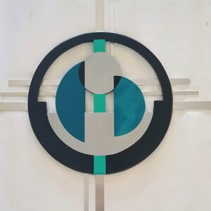 May include: A wall art piece featuring a large black circular frame with horizontal and vertical silver bars extending outward. Inside the circle are two teal semi-circular shapes, a central silver disc, and a vertical teal stripe running through the center.