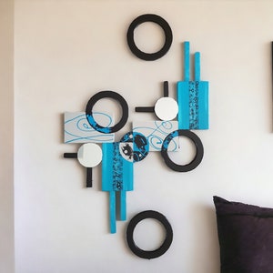 May include: Abstract wall art featuring a geometric design with black circles, white circles, and blue and gray rectangular shapes. The art is mounted on a white wall.