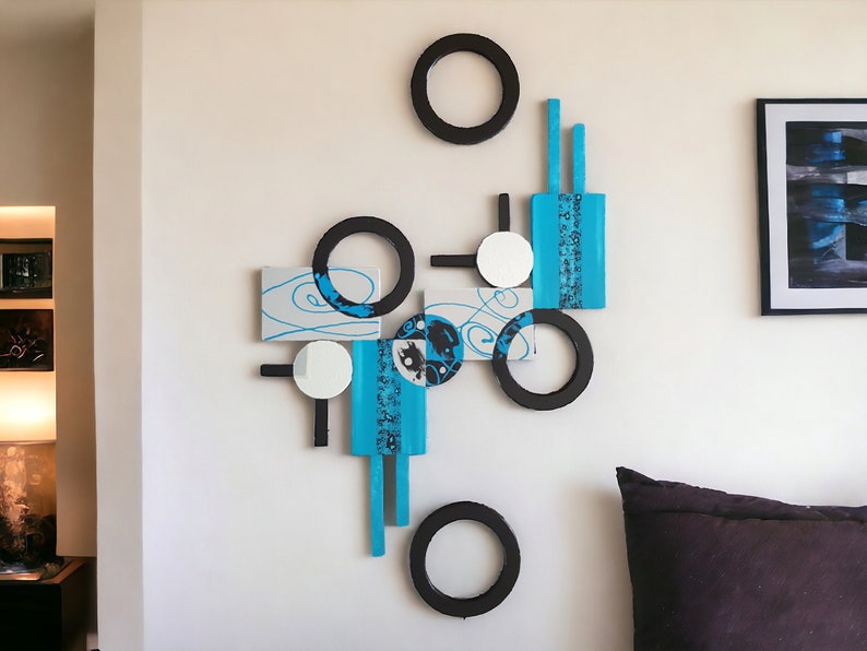 May include: Abstract wall art featuring a geometric design with black circles, white circles, and blue and gray rectangular shapes. The art is mounted on a white wall.