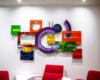 May include: Abstract wall art featuring geometric shapes in various colors, including green, red, orange, yellow, purple, and blue. The shapes are layered and overlapping, creating a dynamic and modern design.