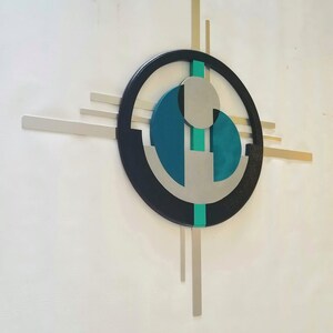 May include: A wall-mounted geometric sculpture featuring a large black circular frame intersected by horizontal and vertical silver metal bars. The center contains a teal circular element, a smaller silver circle, and a vertical teal strip.