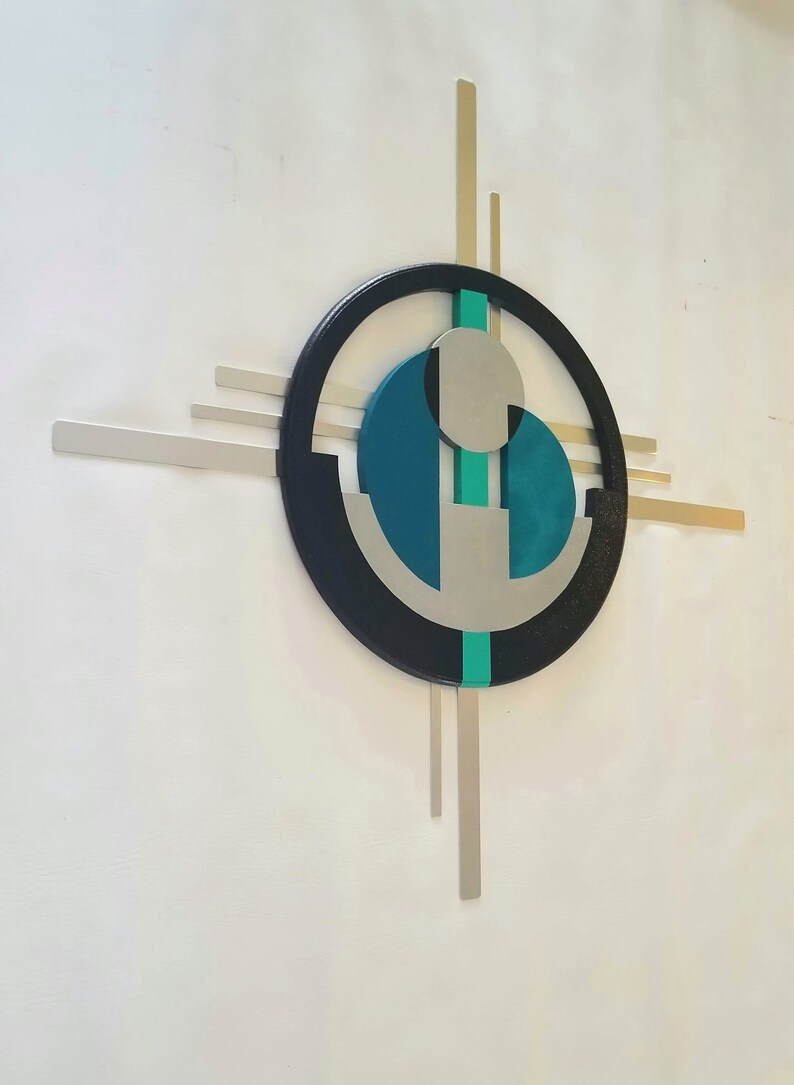 May include: A wall-mounted geometric sculpture featuring a large black circular frame intersected by horizontal and vertical silver metal bars. The center contains a teal circular element, a smaller silver circle, and a vertical teal strip.