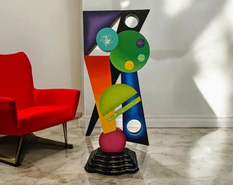May include: Abstract sculpture made of wood and painted in various colors, including green, blue, purple, orange, and pink. The sculpture features geometric shapes and circles, and is mounted on a black base.