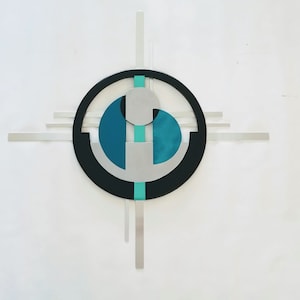 May include: A wall-mounted metal sculpture featuring a central black circle intersected by horizontal and vertical silver bars. The design incorporates teal and silver geometric shapes, including a circular element and two semi-circular forms.