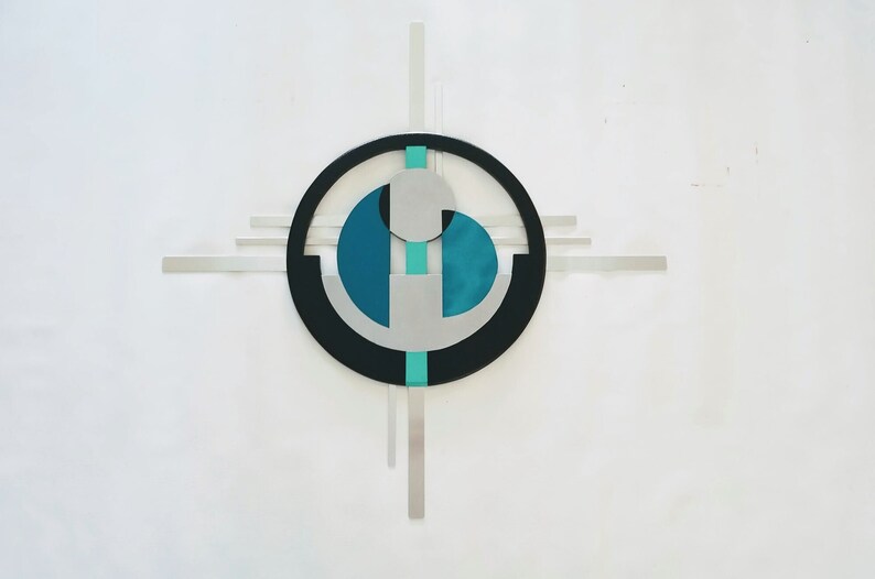 May include: A wall-mounted metal sculpture featuring a central black circle intersected by horizontal and vertical silver bars. The design incorporates teal and silver geometric shapes, including a circular element and two semi-circular forms.