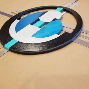May include: A wall art piece featuring a large black oval frame containing layered geometric shapes in white, dark blue, and bright turquoise. Four thin, straight silver metal bars extend horizontally and vertically from behind the oval frame.