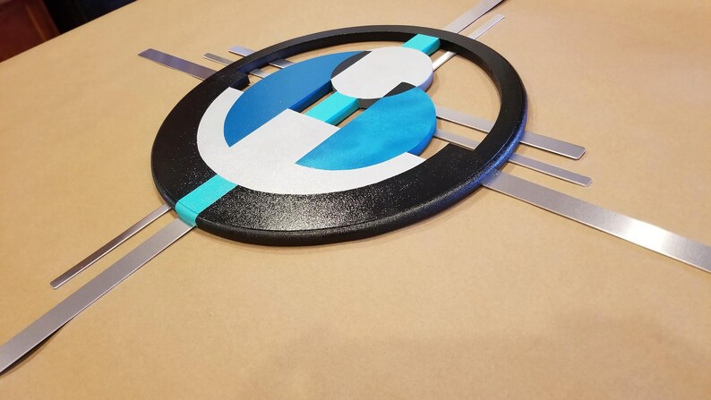 May include: A wall art piece featuring a large black oval frame containing layered geometric shapes in white, dark blue, and bright turquoise. Four thin, straight silver metal bars extend horizontally and vertically from behind the oval frame.