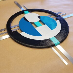 May include: A wall art piece featuring a large black textured circle frame containing geometric shapes in shades of blue, teal, and white. The design is accented by four straight, flat silver metal bars extending outward from behind the central circular frame.