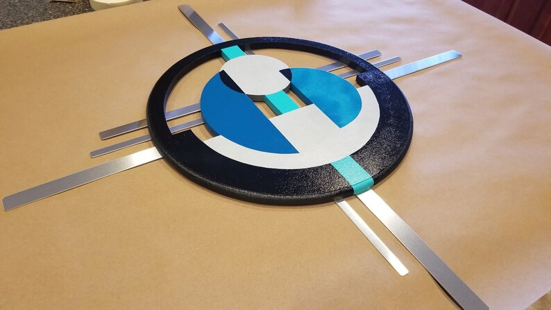 May include: A wall art piece featuring a large black textured circle frame containing geometric shapes in shades of blue, teal, and white. The design is accented by four straight, flat silver metal bars extending outward from behind the central circular frame.