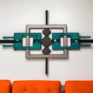 May include: Abstract wall art with a geometric design. The artwork features a teal, gray, and black color scheme. The central design includes squares, rectangles, and diamond shapes, creating a modern aesthetic. The piece is mounted on a white wall.