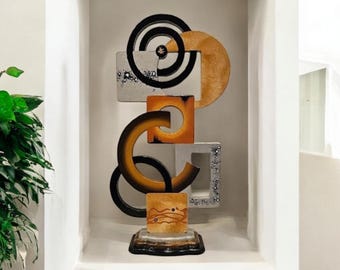 May include: Abstract sculpture made of wood and metal with geometric shapes in shades of gold, silver, and black. The sculpture is on a black base and features a circular design with a smaller circle inside.