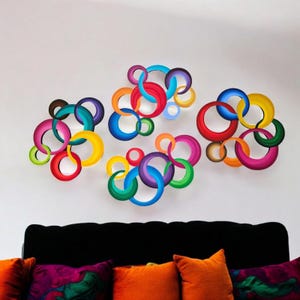 May include: Colorful wall art featuring overlapping, painted rings in various sizes and hues, including red, blue, yellow, and green. The abstract design is mounted on a white wall above a black sofa with orange and patterned pillows.