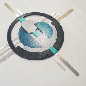 May include: A wall art piece featuring a large black circular frame with a textured finish. Inside the circle are layered geometric shapes in silver, teal, and dark blue. Four thin, straight silver metal bars extend outward from the center.