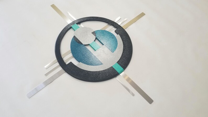 May include: A wall art piece featuring a large black circular frame with a textured finish. Inside the circle are layered geometric shapes in silver, teal, and dark blue. Four thin, straight silver metal bars extend outward from the center.