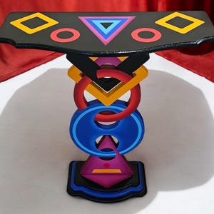 May include: A black console table featuring a top surface decorated with yellow squares, red circles, and a central blue and purple triangle. The pedestal base consists of stacked, interlocking geometric shapes in red, yellow, blue, and purple.