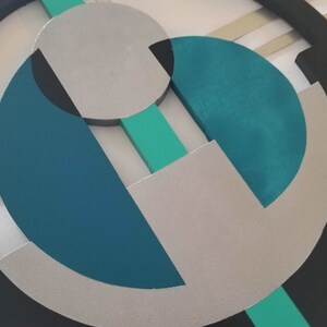 May include: A circular wall art piece featuring a black outer ring, a central silver circle, and geometric shapes in teal, dark blue, and mint green. The composition includes intersecting lines and layered circular segments arranged in a balanced pattern.
