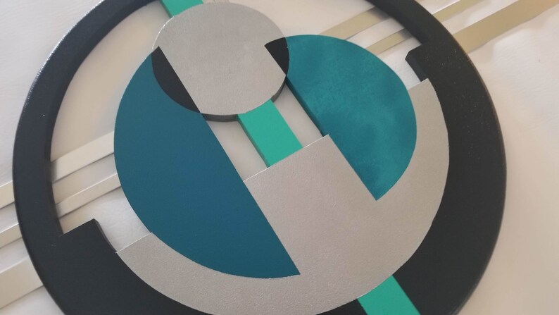 May include: A circular wall art piece featuring a black outer ring, a central silver circle, and geometric shapes in teal, dark blue, and mint green. The composition includes intersecting lines and layered circular segments arranged in a balanced pattern.