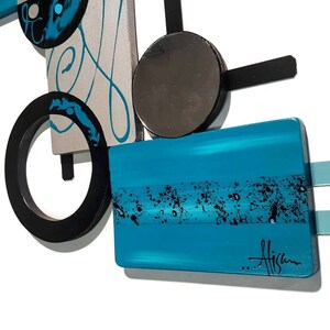 May include: Abstract wall art with geometric shapes in black, silver, and turquoise blue. The piece features a circle, a rectangle, and a square, with a mirrored circle in the center. The artist's signature is visible in the lower right corner.