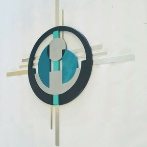 May include: A wall sculpture featuring a large black circular frame with intersecting horizontal and vertical silver metal bars. The center contains a teal vertical strip, two teal semi-circular shapes, and a light gray circular element.